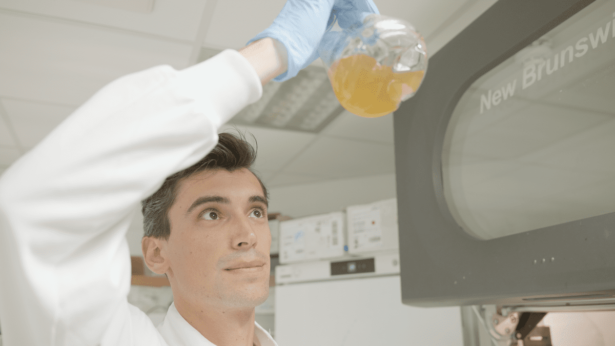 A young, maybe 20-year-old man in white clothing and plastic gloves holds a transparent vessel, similar to a test tube, in front of him and above his head, while he gazes at it