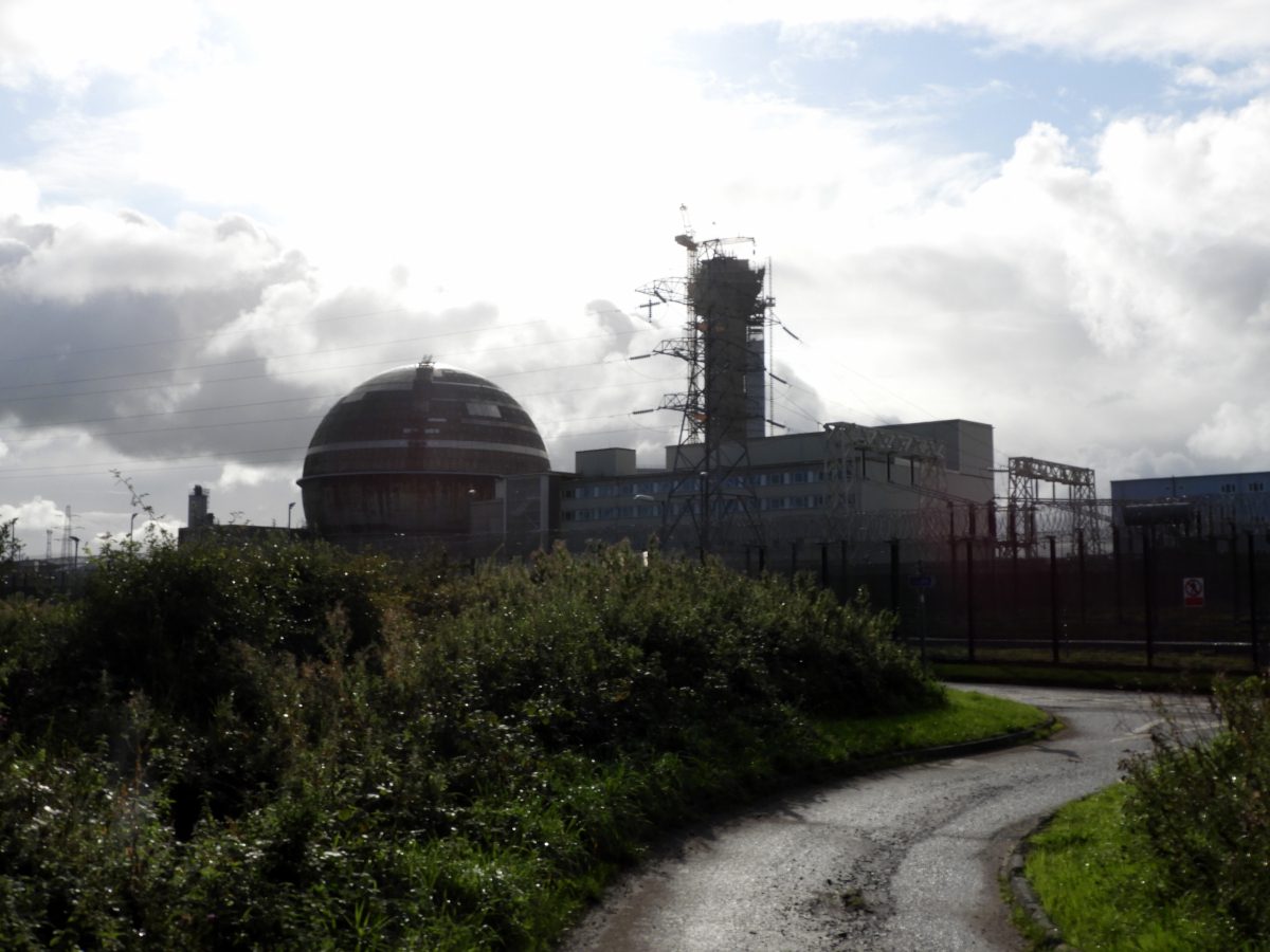 A collection of industrial buildings, chimneys and other structures on a flat, green, coastal expanse of land with a hill or elevated ridge visible in the far distance