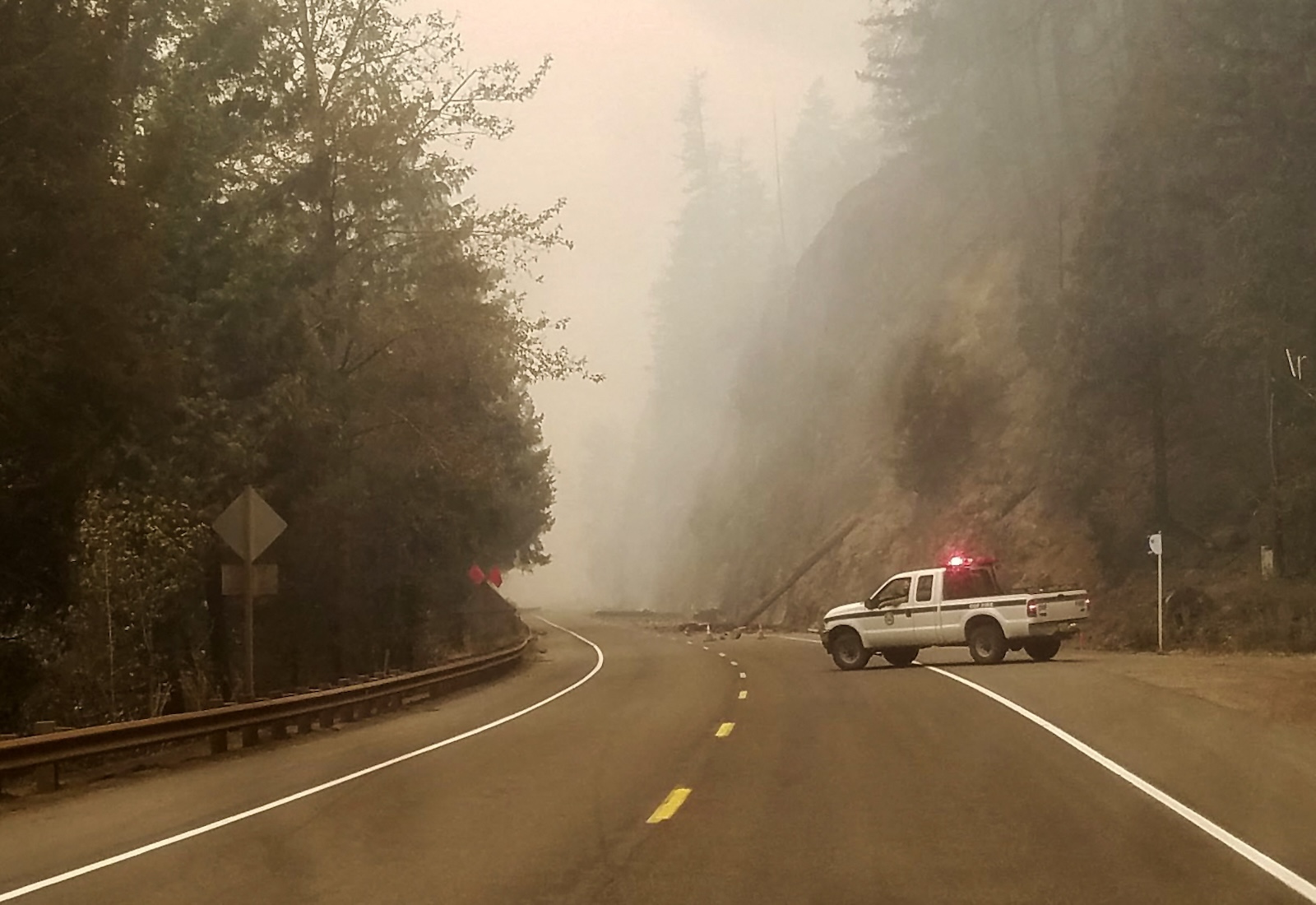 Road with emergency vehicle parked on the right side, ahead of a felled tree. Forests are on either side of the road, and the air is smoky.